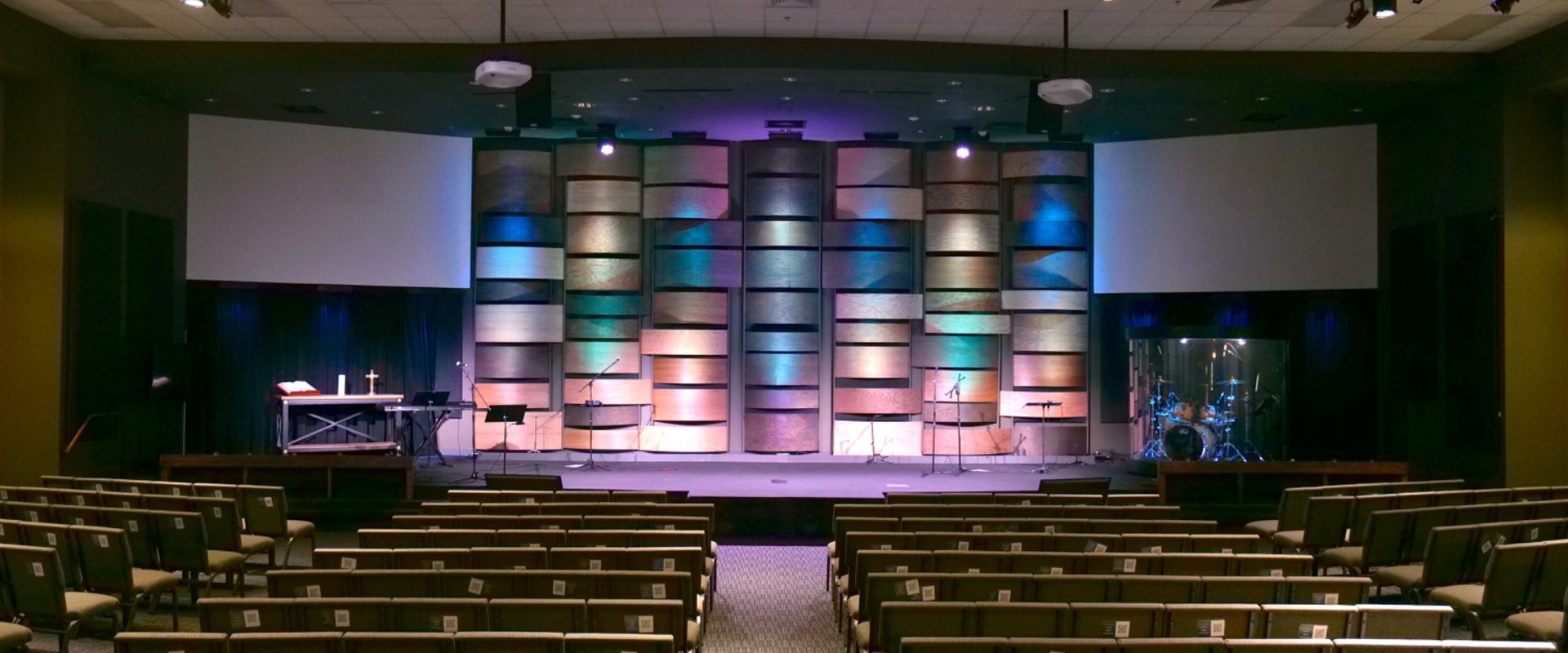 Modern church sanctuary interior with contemporary wooden stage backdrop, colorful LED lighting, professional worship setup, and rows of wooden pews